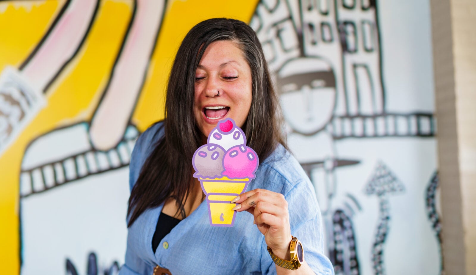 A woman holding up a die-cut flyer in the shape of an ice cream cone, pretending to take a bite out of it.