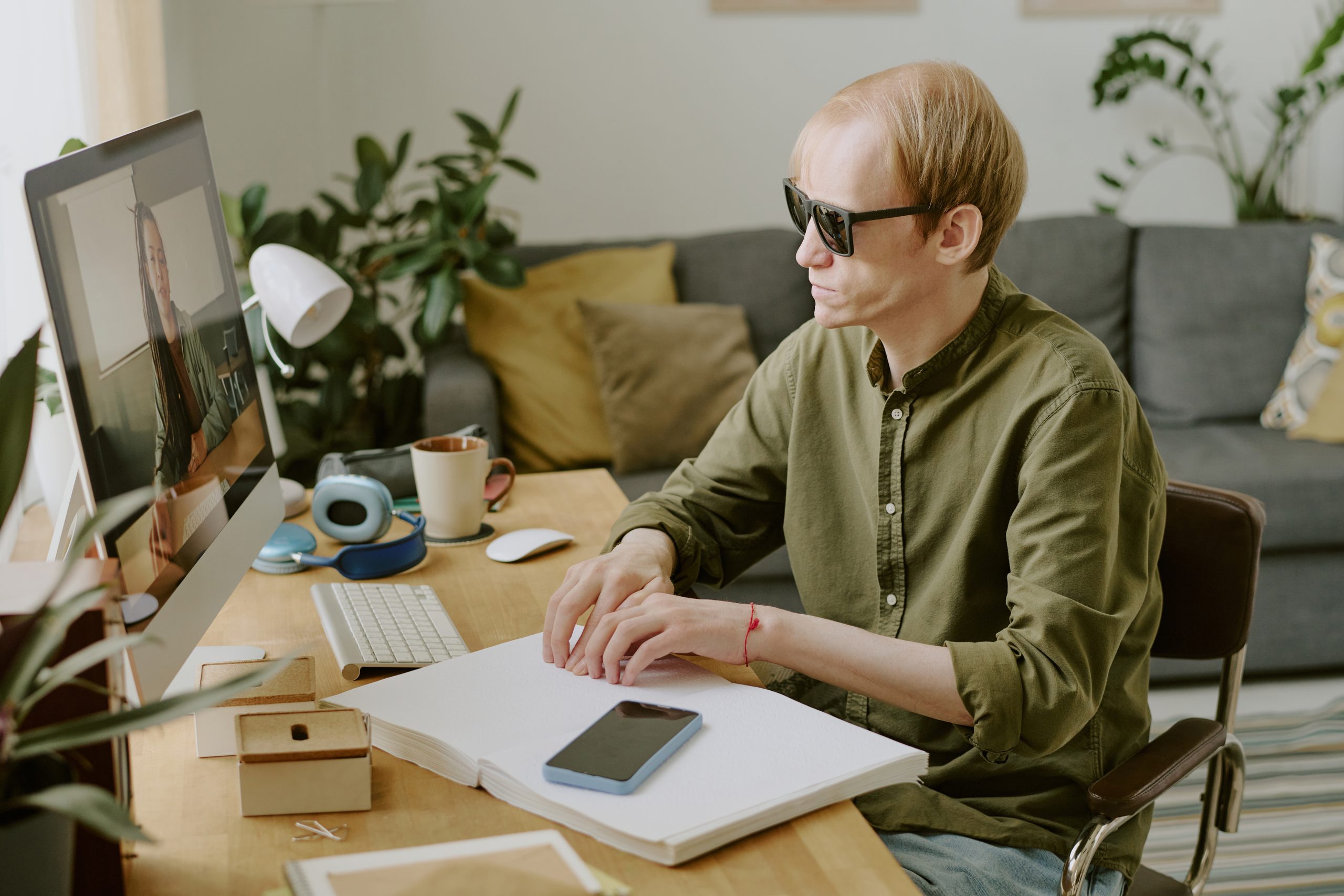 A caucasian man wearing sunglasses sitting at a computer desk with his hands reading an open braille book.