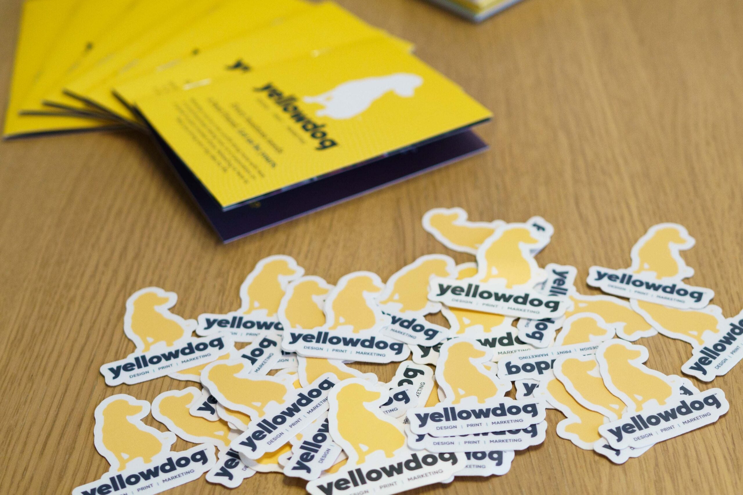 Cluster of YellowDog branded stickers on a wooden table. Small yellow booklet/brochures and a YellowDog recycled notebook are visible in the background.